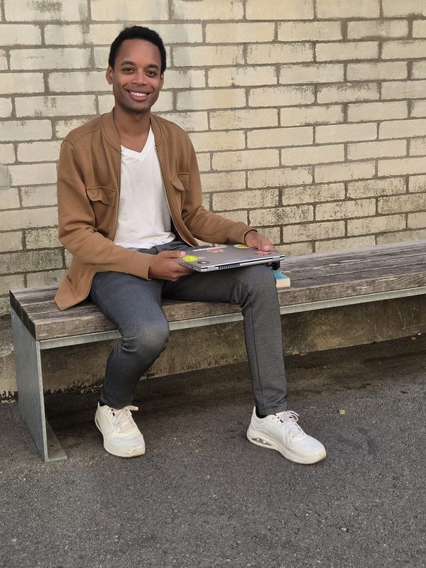 Fabrice Brunner sitzt mit seinem Laptop auf einer Bank und lächelt in die Kamera.