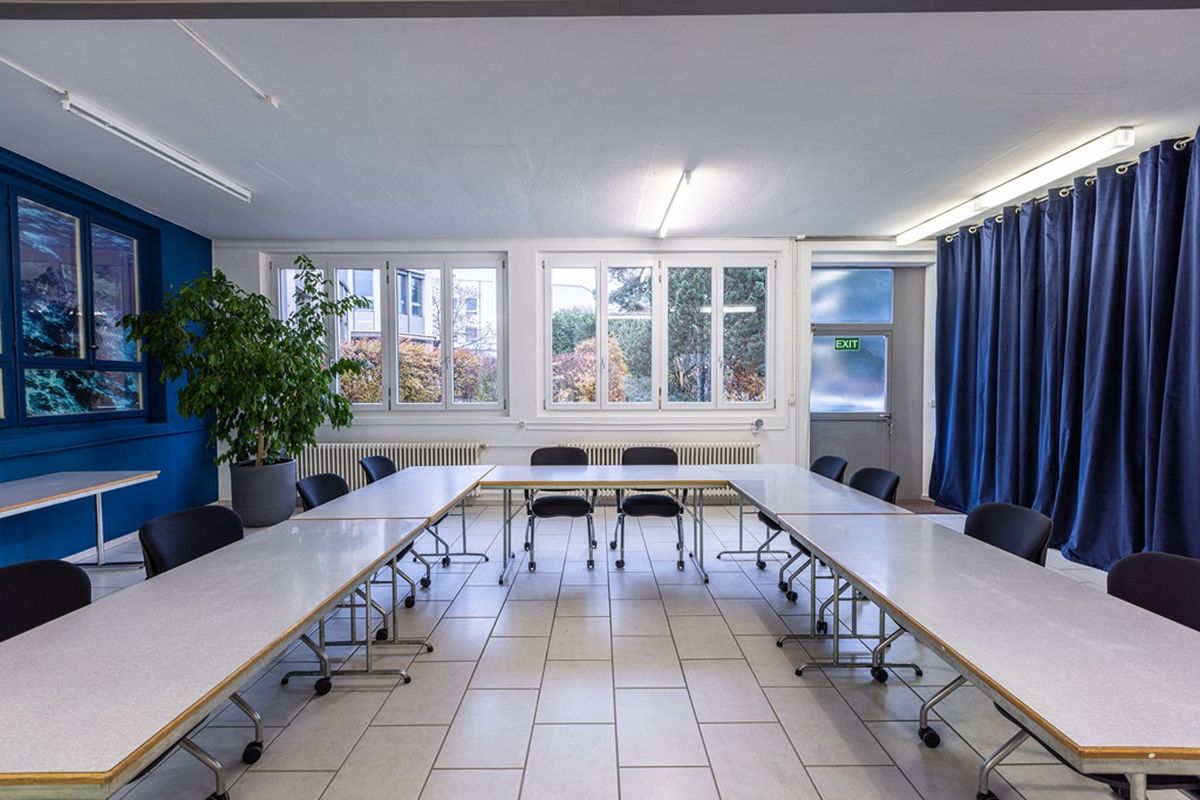 Derselbe Kursraum mit langer Tischreihe, Stühlen und Pflanze vor der blauen Wand.